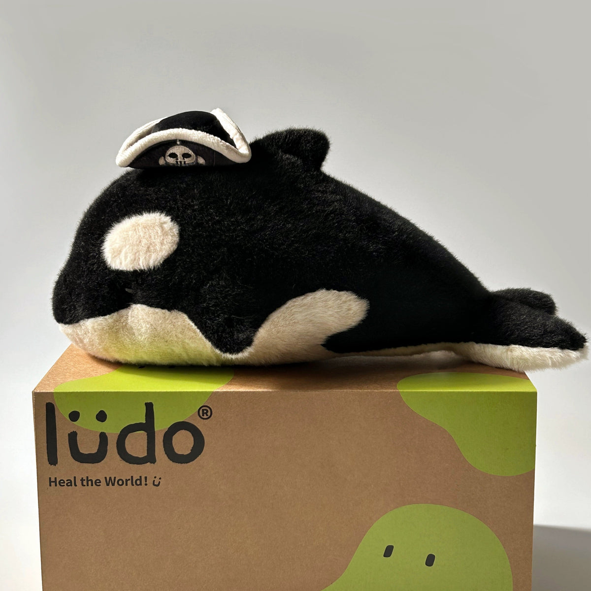 A plush toy resembling an orca whale, with black and white coloring, sitting on a box with the brand name 'lüd' on it.