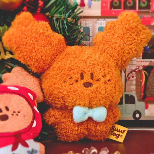 Plush toy with a bow tie in front of a Christmas tree with ornaments and a toy bus.