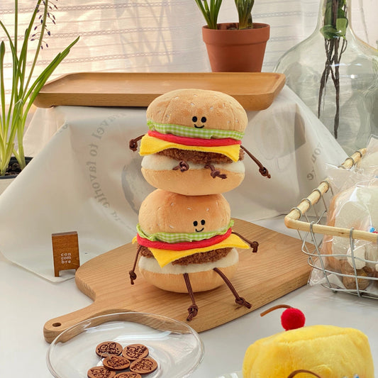 Plush toy hamburger on a wooden board with plants and a cake in the background