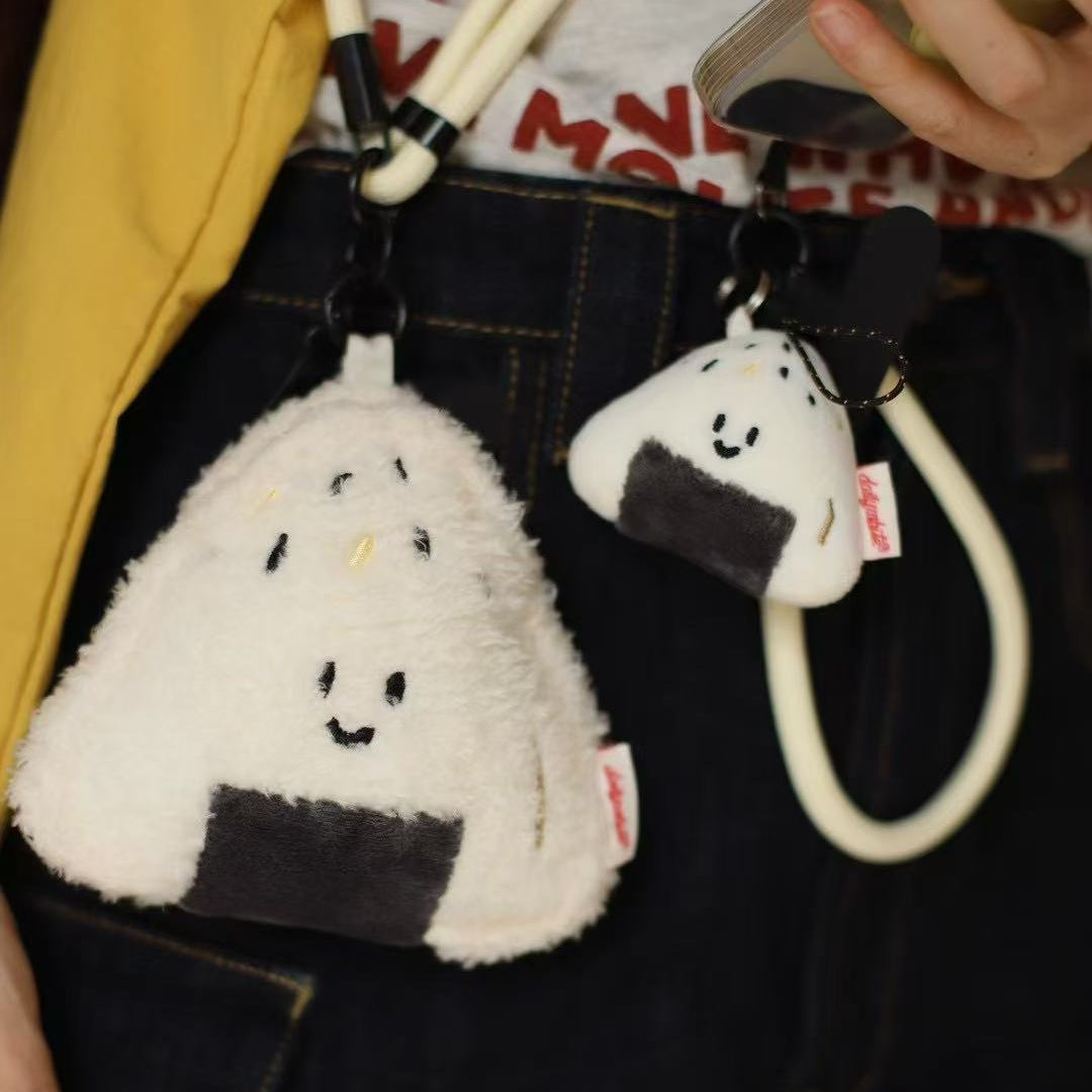 Two plush rice ball keychains hanging from a strap, with a person holding a phone in the background.