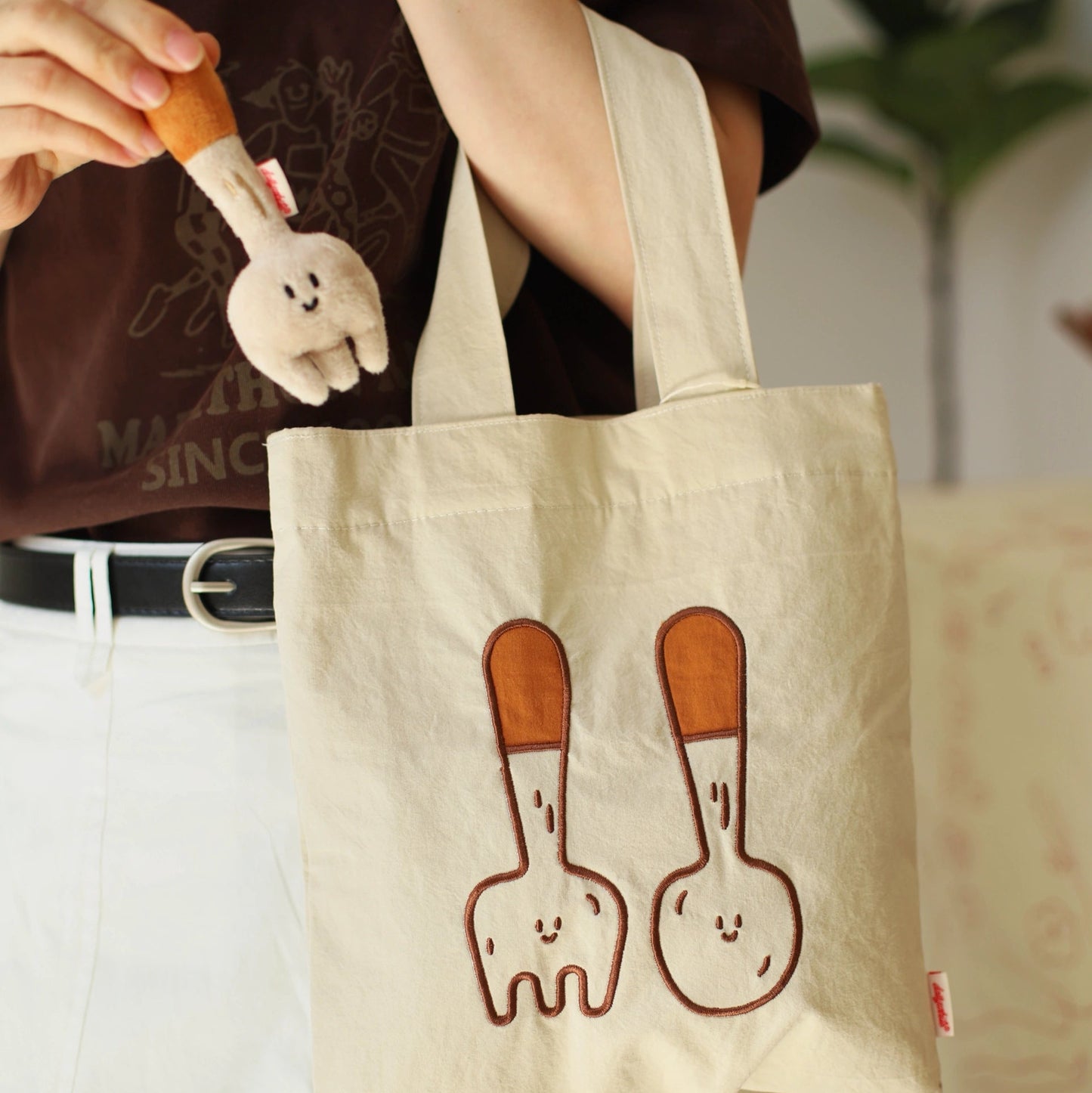 Person holding a tote bag with cartoon designs of a fork and spoon, wearing a brown shirt and white pants.