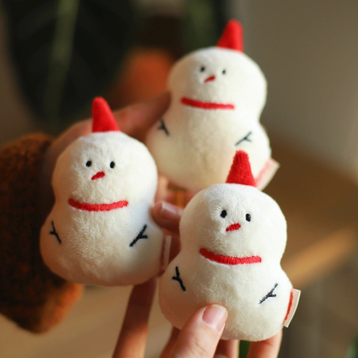 Three small plush chicken toys with red hats held in a hand against a blurred background.