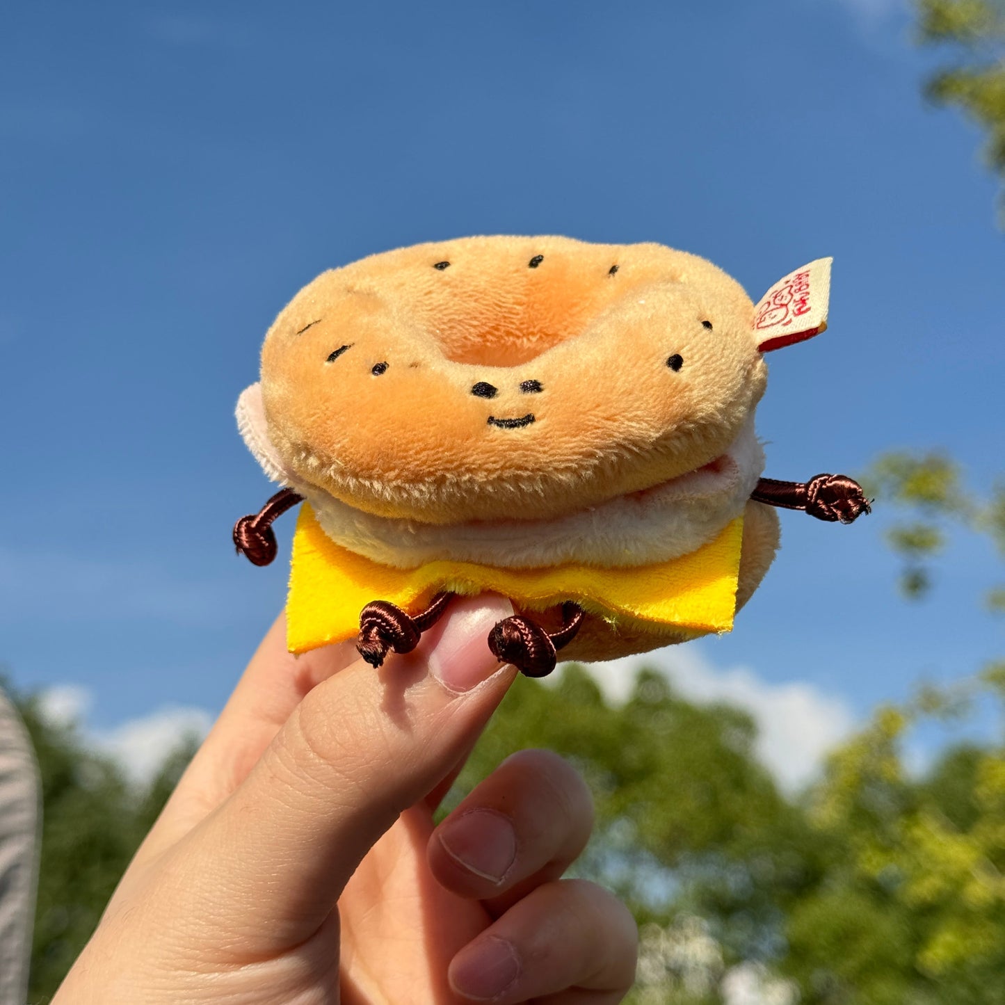 Plush toy resembling a sandwich held by a hand against a blue sky with trees in the background