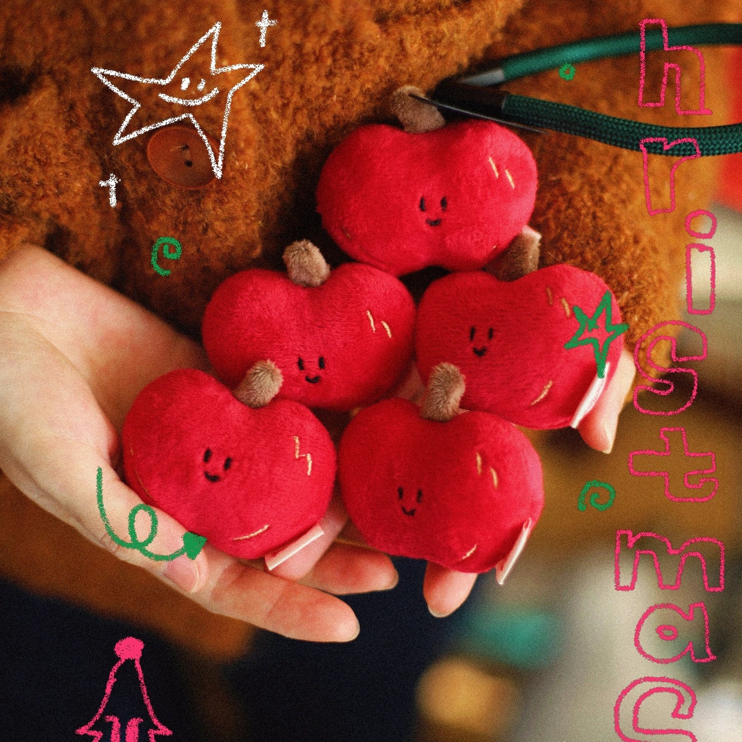 Red plush apples with smiling faces held by a hand, surrounded by festive text and decorations.