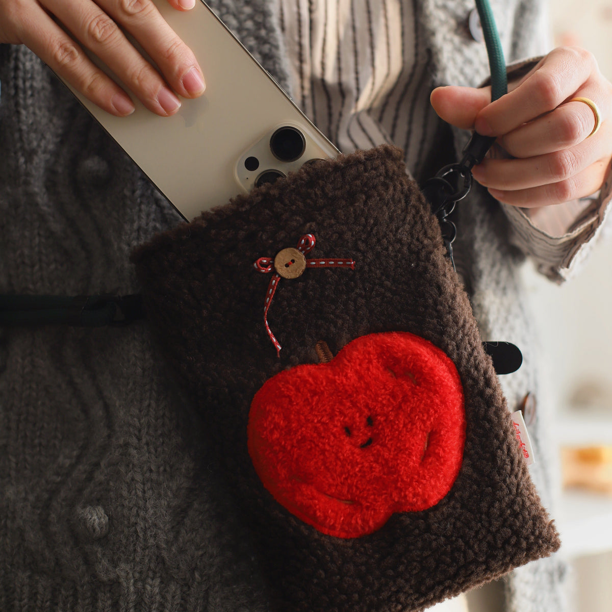 Person holding a brown pouch with a red apple design and a phone, wearing a gray sweater.