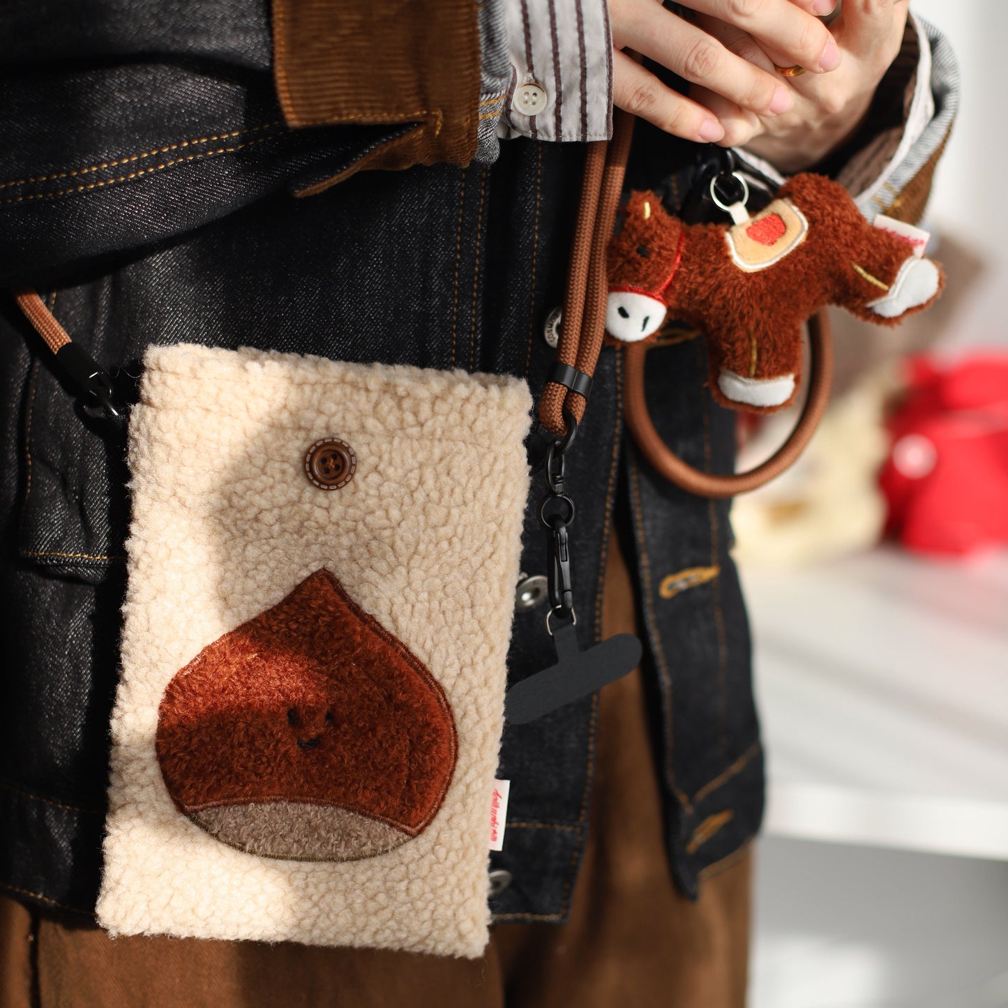 Person holding a phone with a plush toy attached, wearing a dark jacket with a fur-lined pocket.