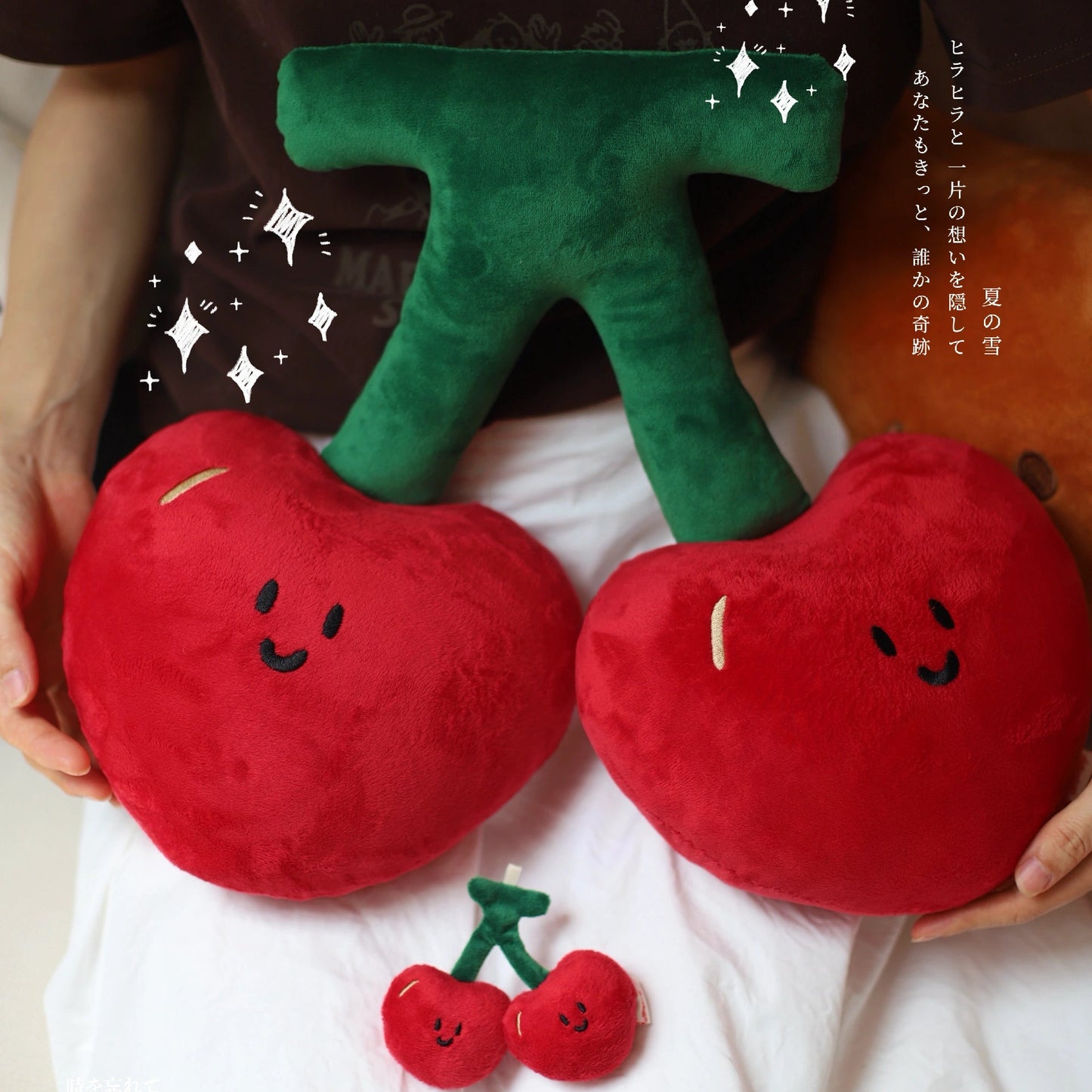 Plush toys shaped like cherries and a leaf held by hands on a white surface.
