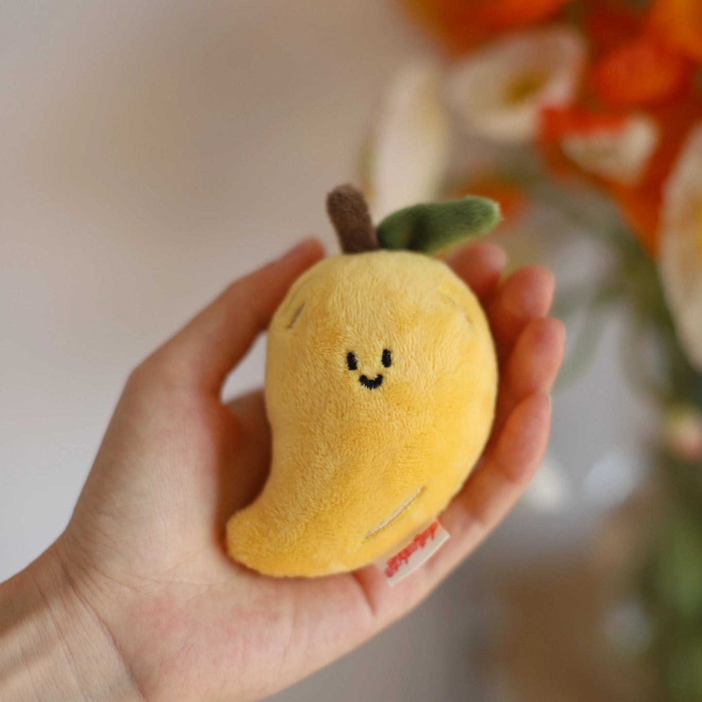 Hand holding a plush mango toy with a blurred floral background
