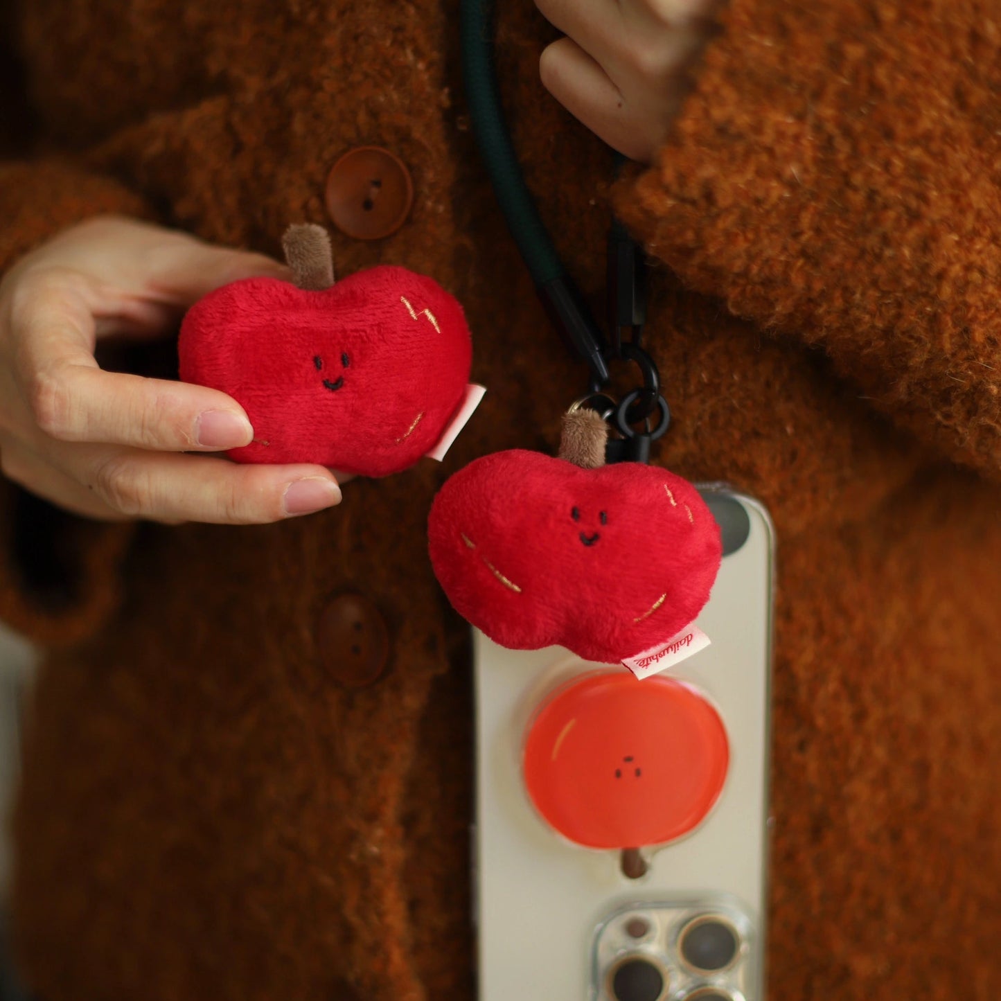 Red apple-shaped phone charm attached to a phone case, held by a person wearing a brown coat.