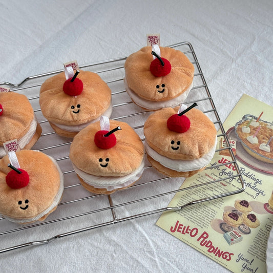 Plush toy pastries with cherry on top on a cooling rack next to a vintage Jello Puddings recipe card.