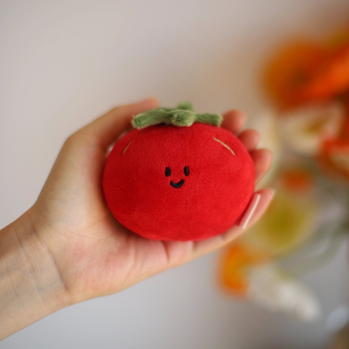 Hand holding a plush toy shaped like a red tomato with a smiling face against a blurred background.