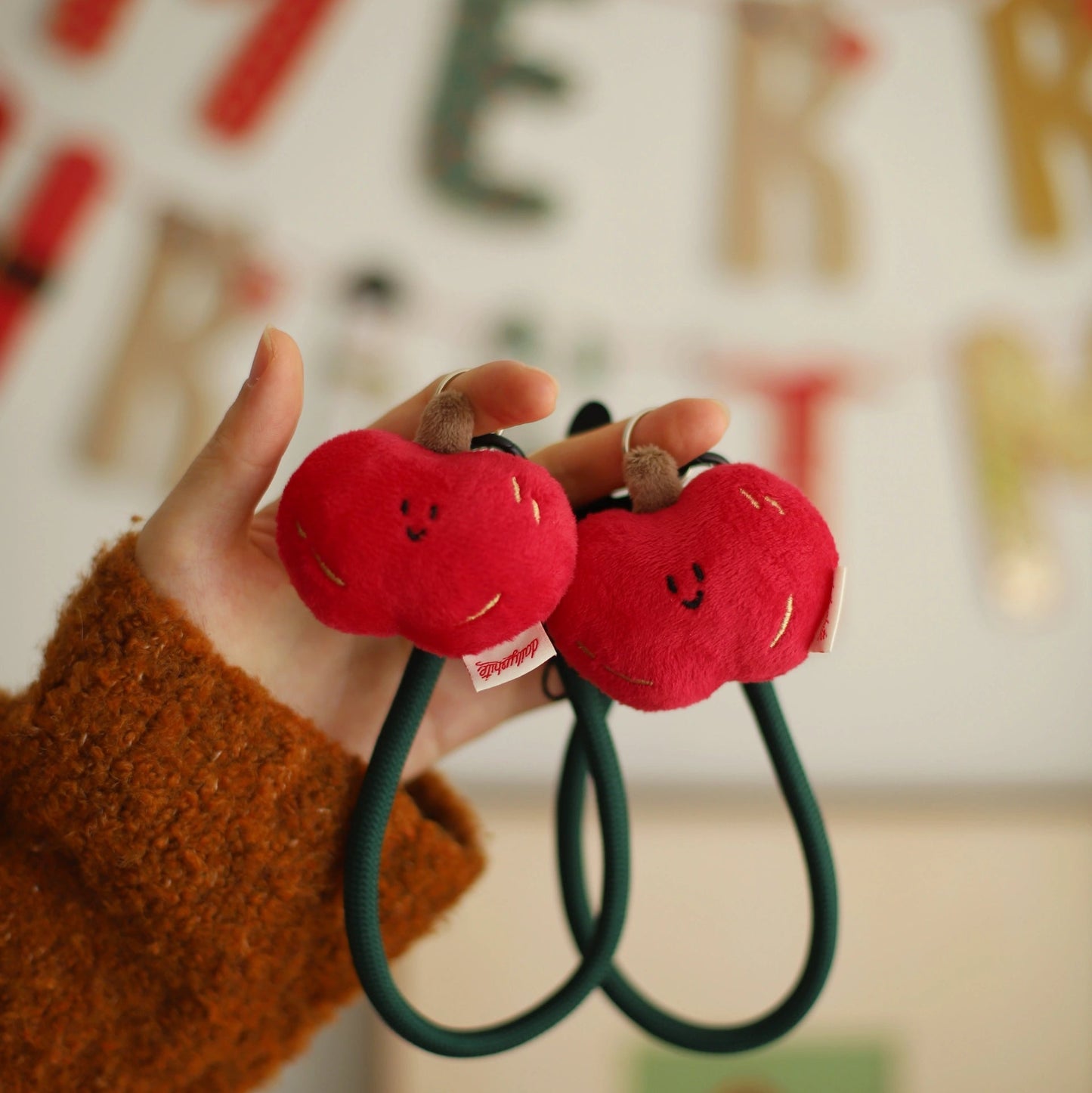 Red plush apple-shaped hair ties held by a hand with a festive background.