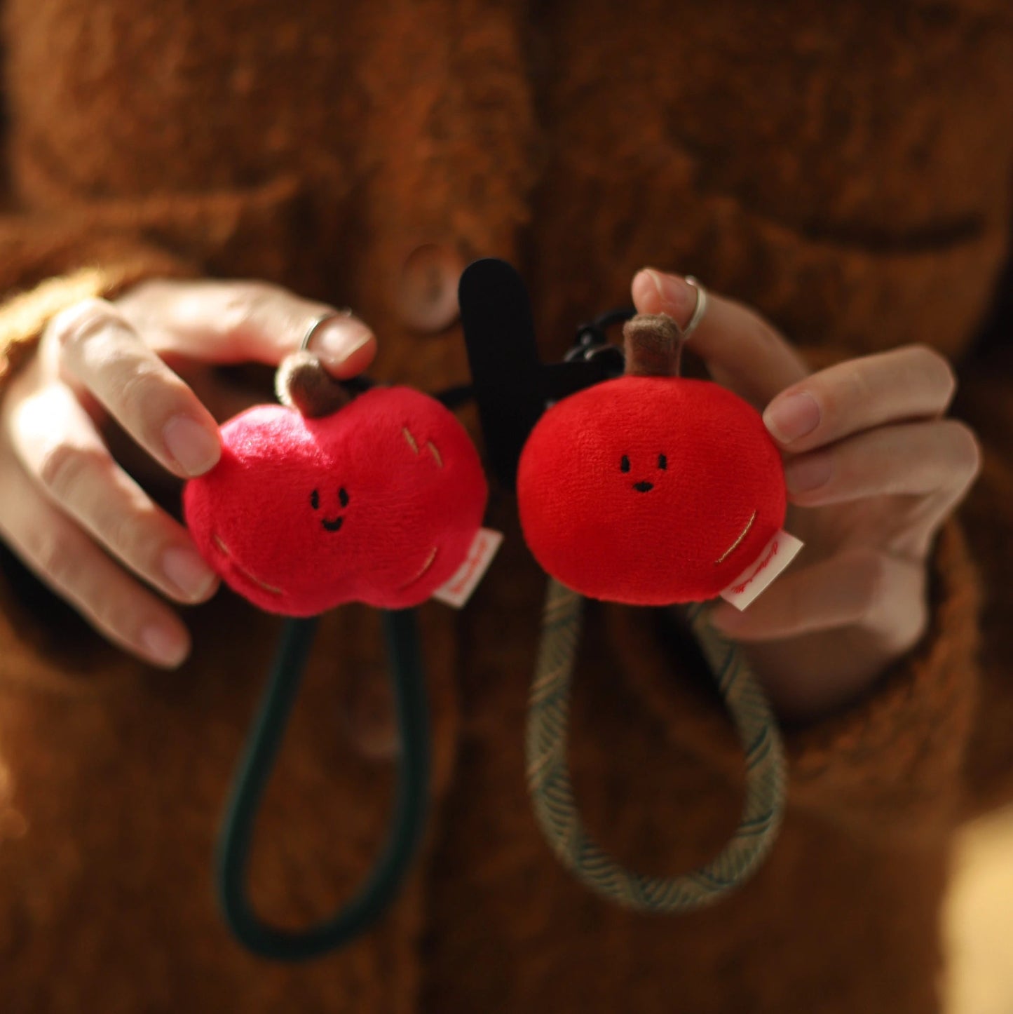 Red plush keychains shaped like apples held by a person wearing a brown coat.