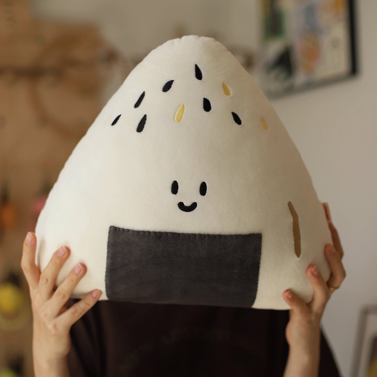 Person holding a plush rice ball toy with a smiley face in a room setting.