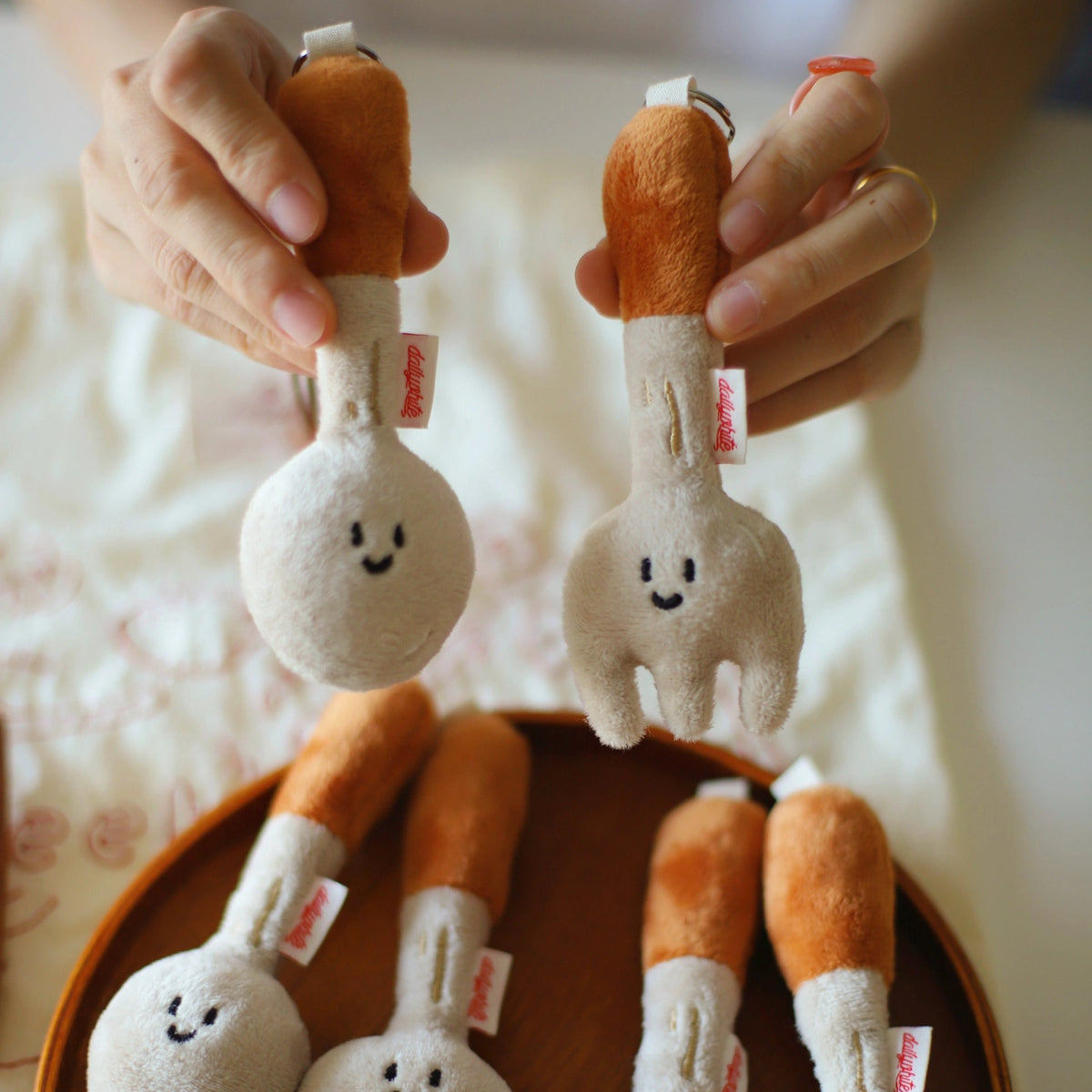 Plush spoon and fork keychains with smiling faces held by hands above a wooden surface.