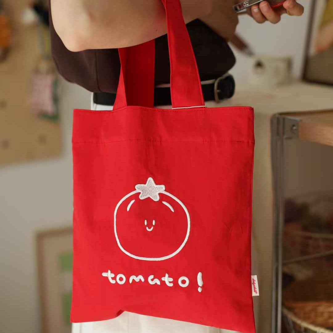 Red tote bag with a tomato design and text held by a person.