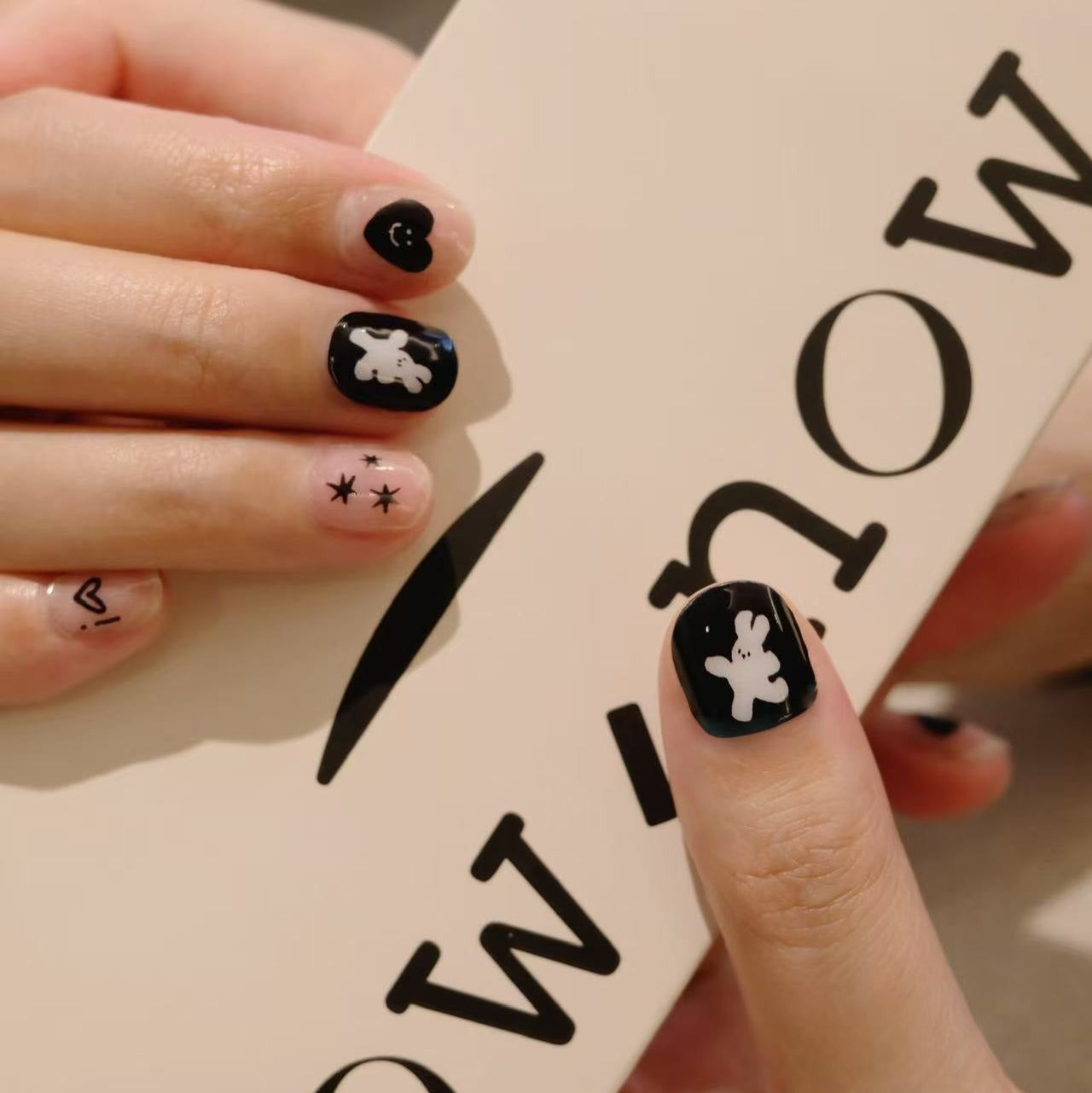 Person holding a box with 'NOW' text, showcasing black nail art with white designs.
