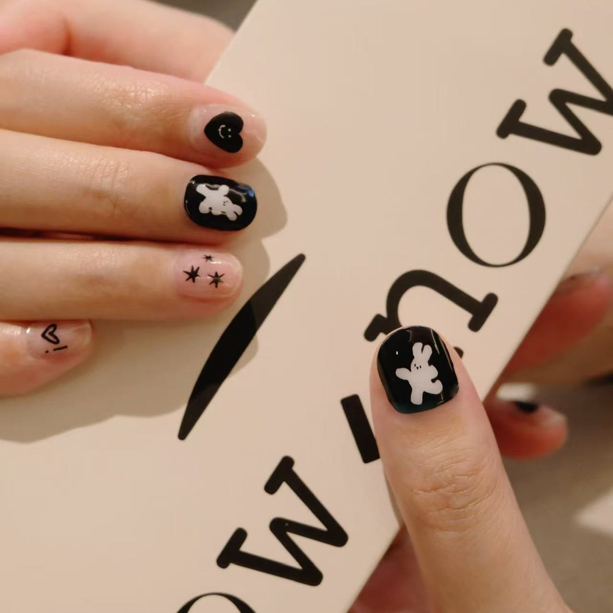 Person holding a box with 'NOW' text, showcasing black nail art with white designs.