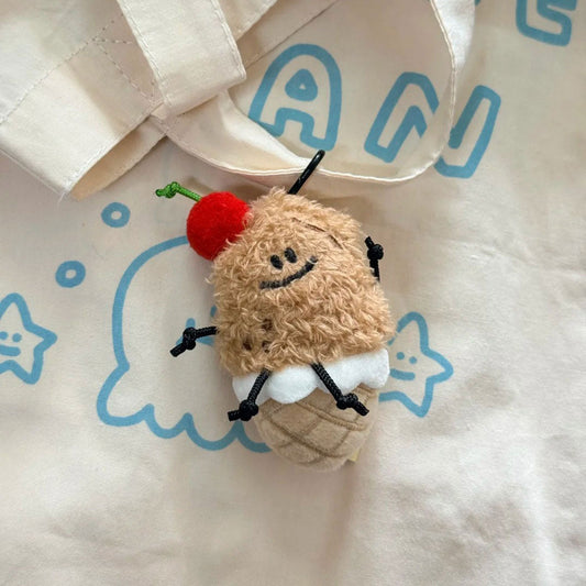 A small plush character charm shaped like a cupcake with a red cherry on top, resting on a cream tote bag with blue cartoon graphics.