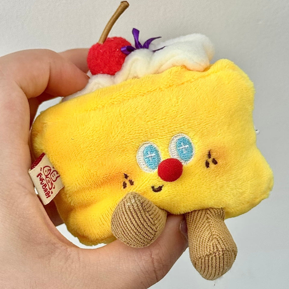 Plush toy resembling a slice of cake with a cherry on top held in a hand against a plain background