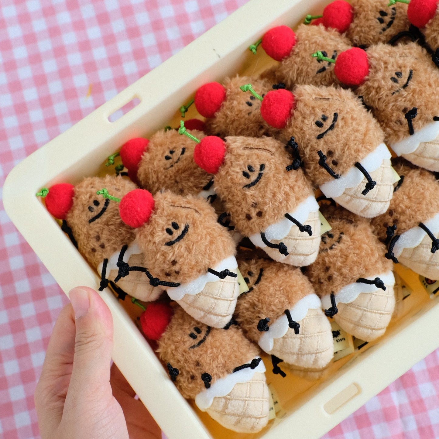 A hand holds a beige plastic tray filled with small plush toy characters dressed in white and beige outfits, each with a red pom-pom on top, set against a pink and white checkered background.