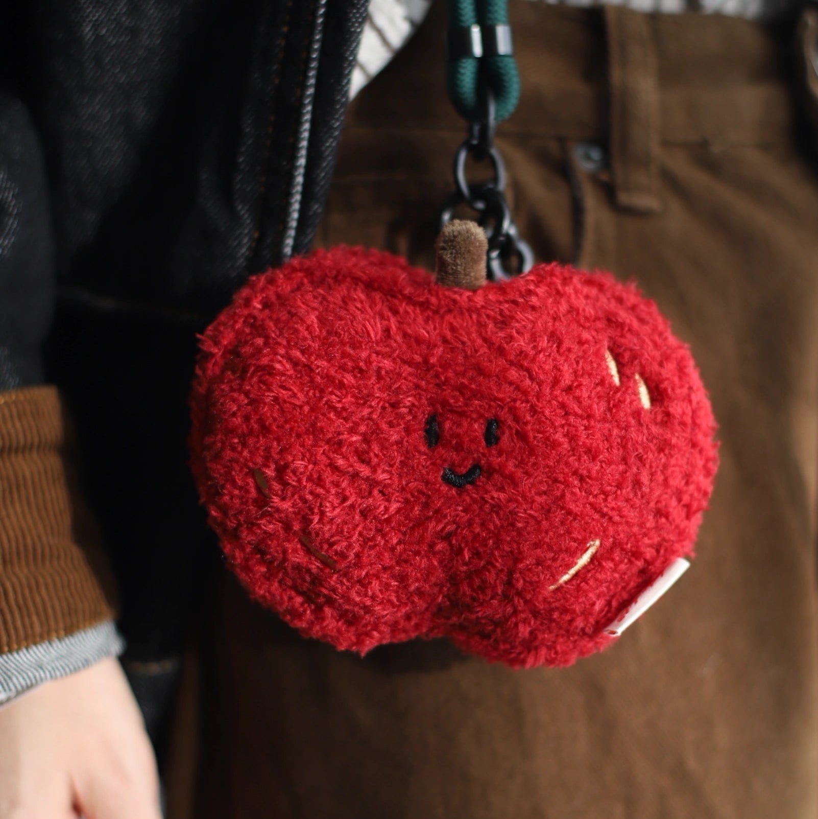 Red plush keychain shaped like a apple held by a person wearing a brown coat.