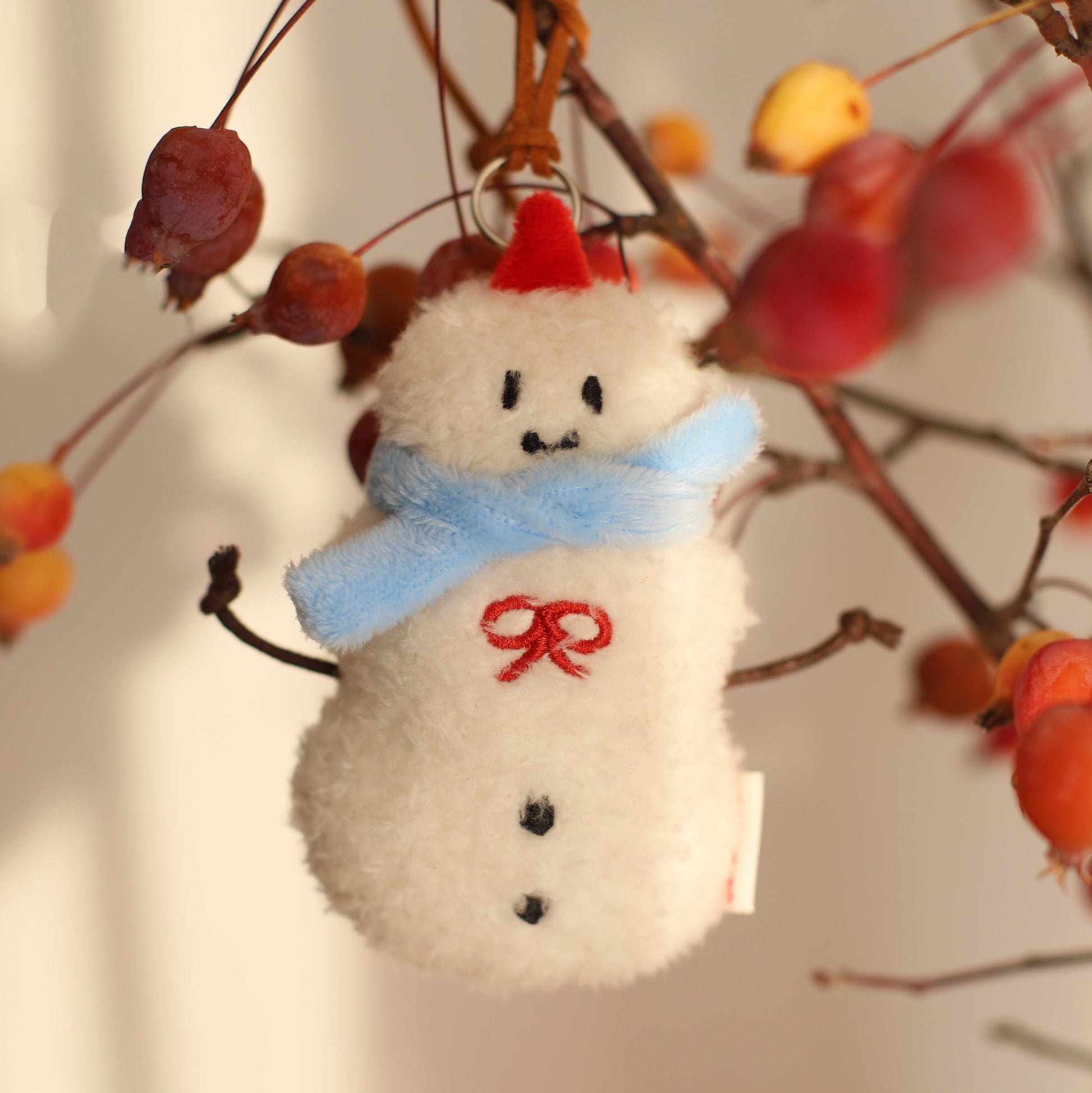 Fuzzy snowman ornament with a red hat and blue scarf hanging on a branch with berries.