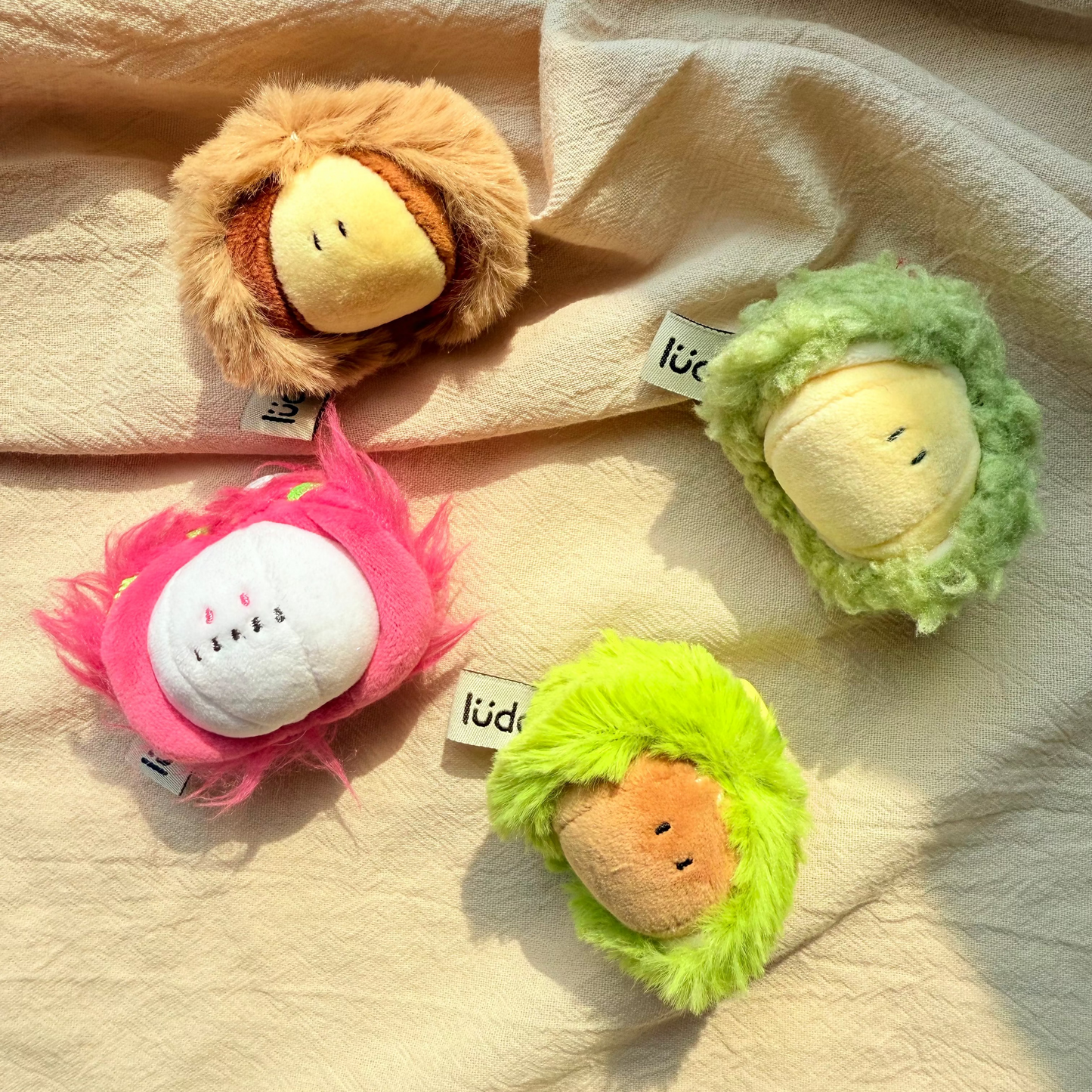 Four small plush character toys arranged on a beige fabric background. The toys have fuzzy textured outer layers in tan, green, pink, and lime green, with round pale yellow faces.
