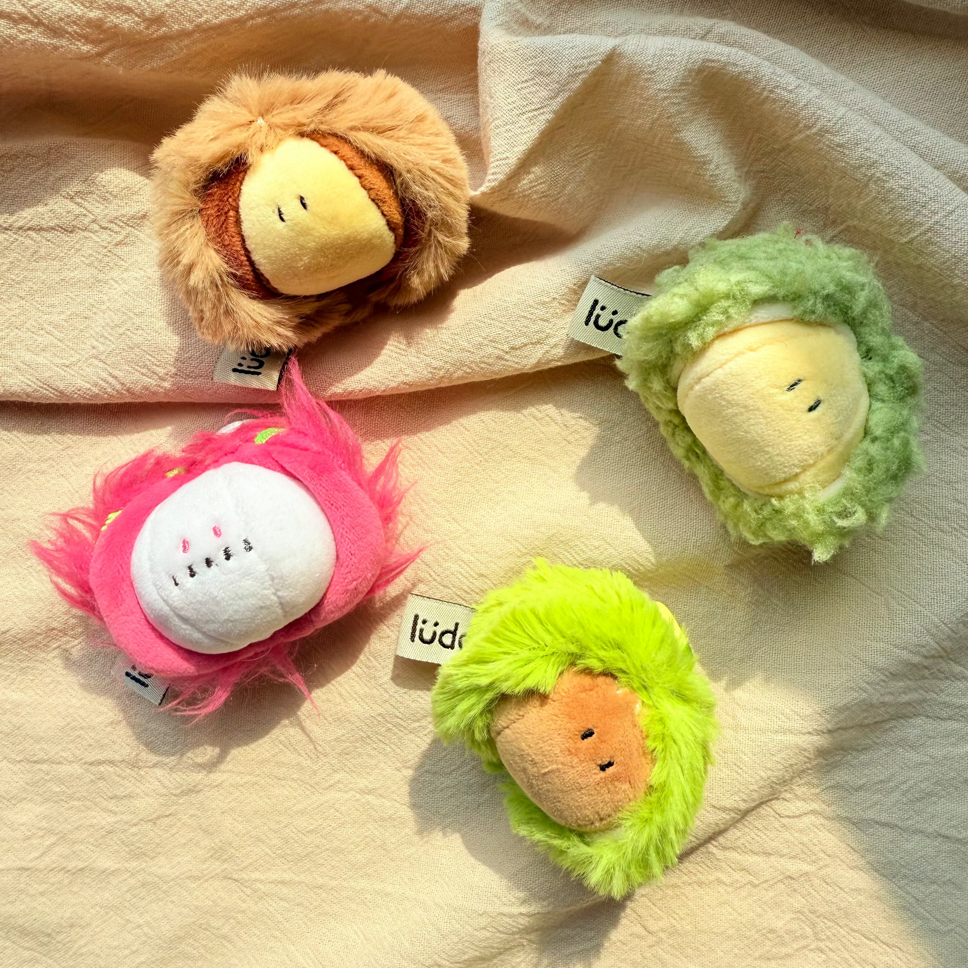 Four small plush character toys arranged on a beige fabric background. The toys have fuzzy textured outer layers in tan, green, pink, and lime green, with round pale yellow faces.