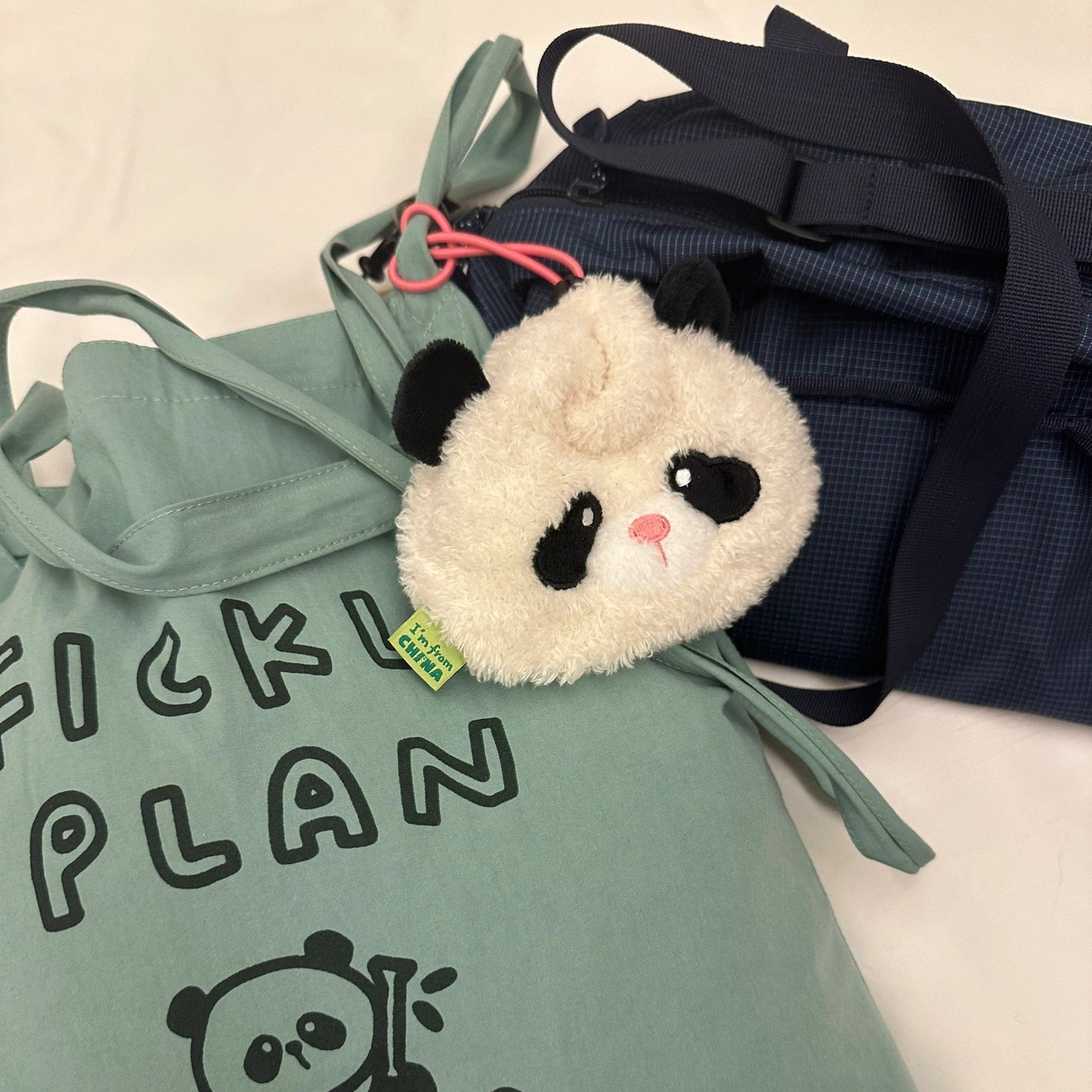 Light green drawstring bag with panda design, plush panda toy, and navy blue bag on a light background