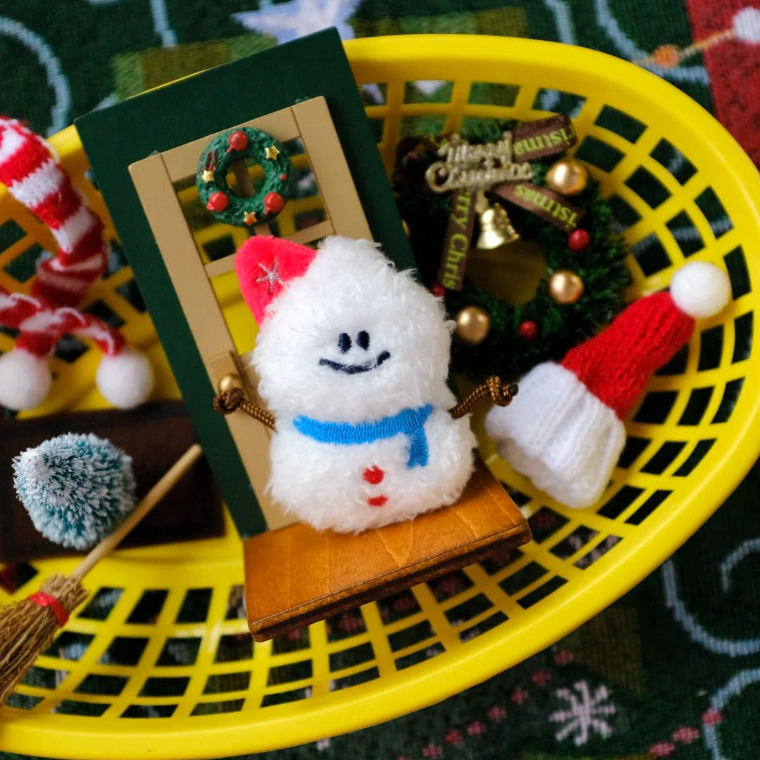Small plush snowman toy with a green door on a decorative surface with Christmas-themed items.