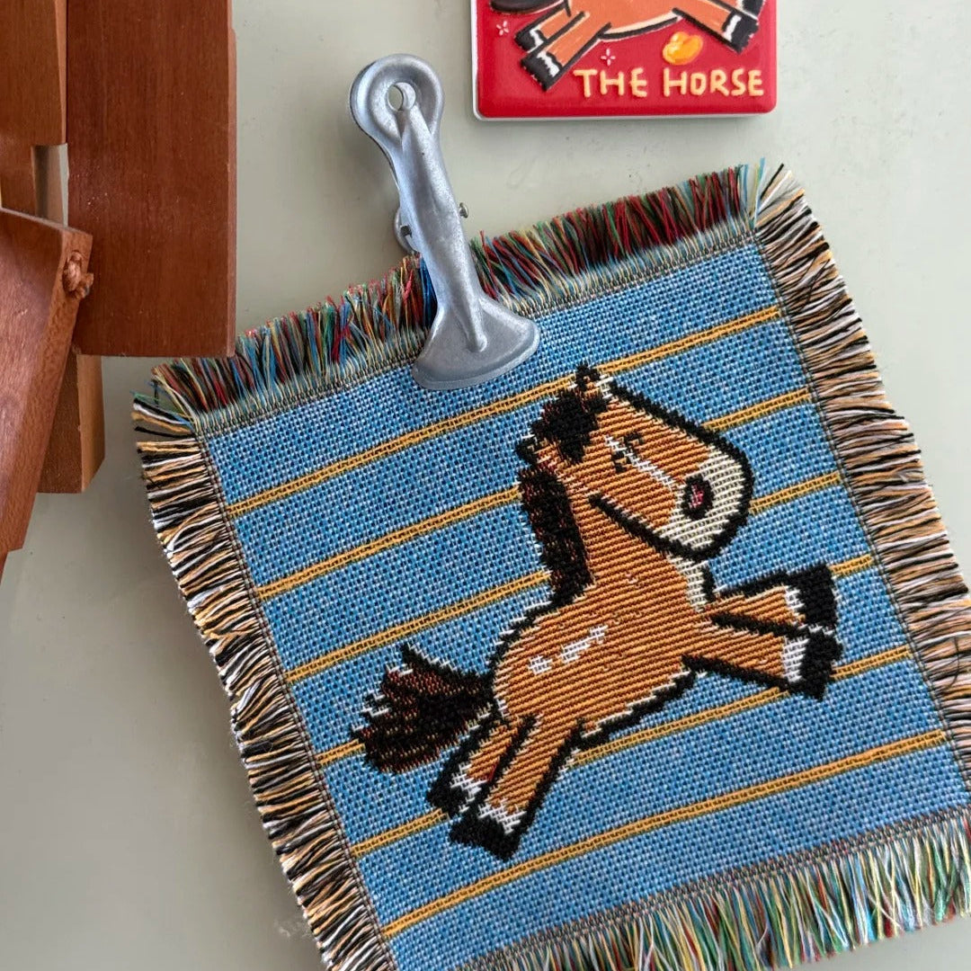 Beaded horse blanket with a metal clip on a white surface, with a small red card featuring a horse illustration in the background.