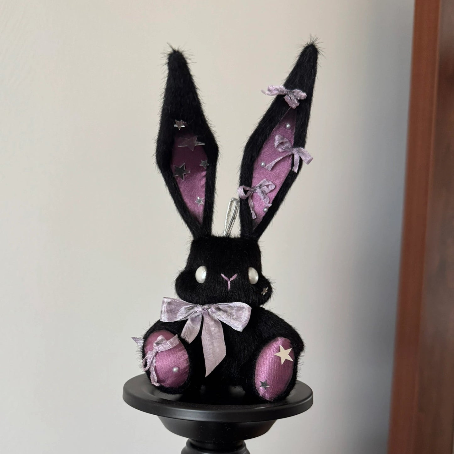 Black rabbit plush toy with pink details on a black stand against a white background