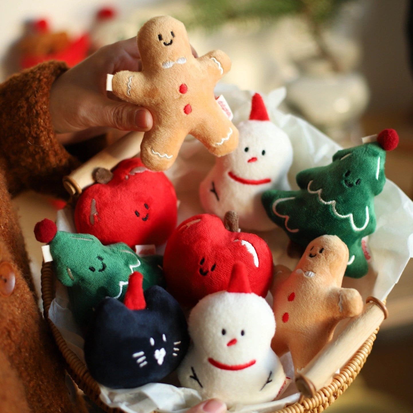 Hand holding a plush gingerbread man toy with other Christmas-themed plush toys in a basket.