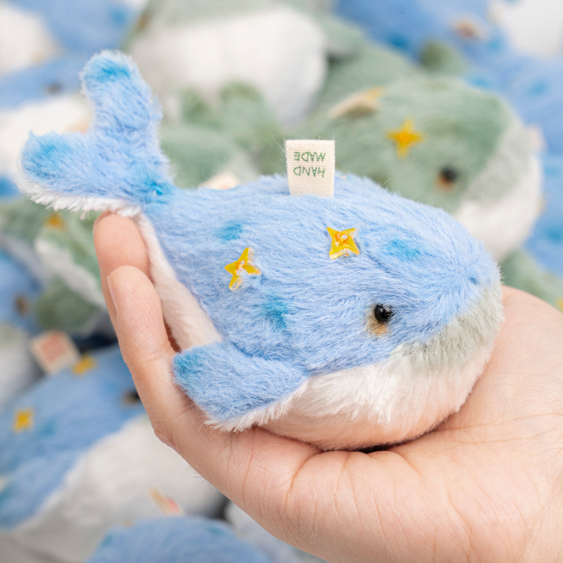 Hand holding a blue plush whale toy with a blurred background of similar toys.