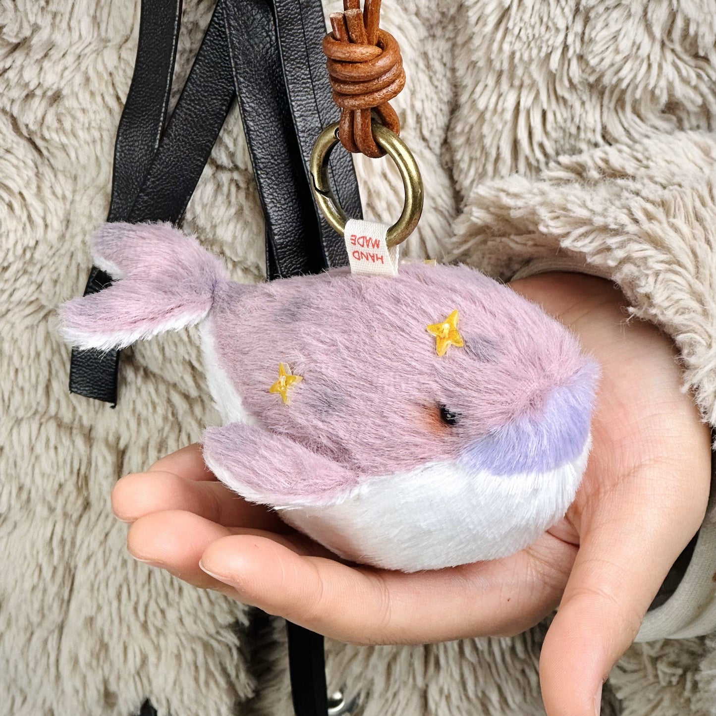 Plush whale toy held in a hand with a strap and ring, on a fluffy surface.