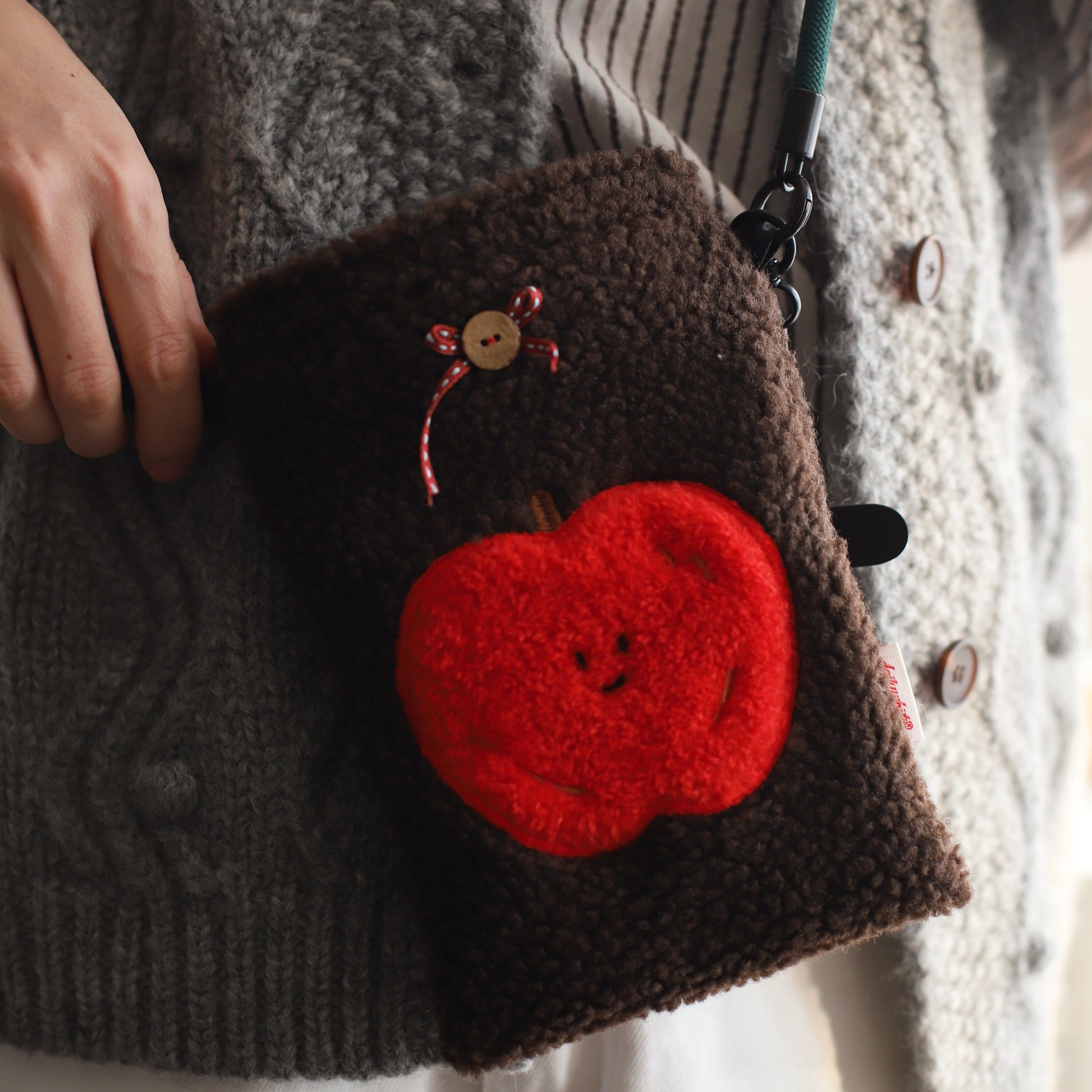 Brown clutch bag with a red apple design held by a person wearing a textured sweater.