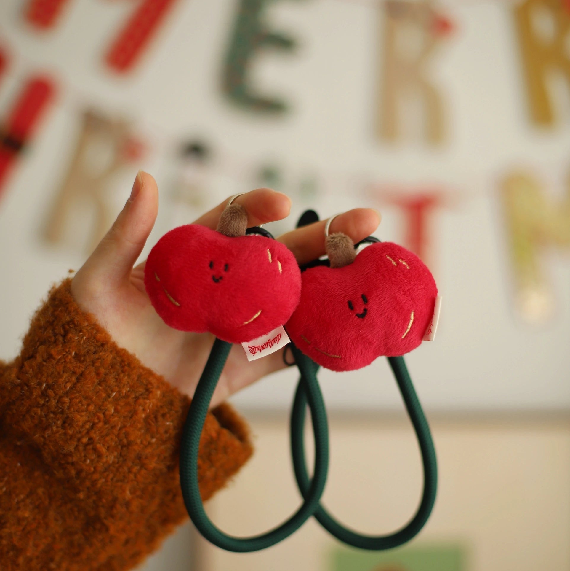 Red plush apple-shaped hair ties held by a hand with a festive background.