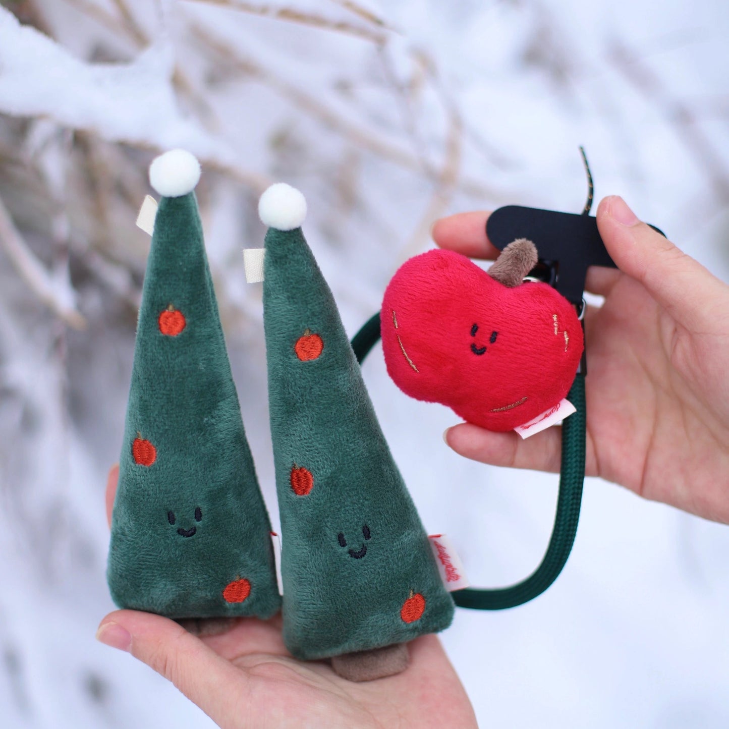 Two green plush trees with red dots and a red plush apple held by a hand against a snowy background.