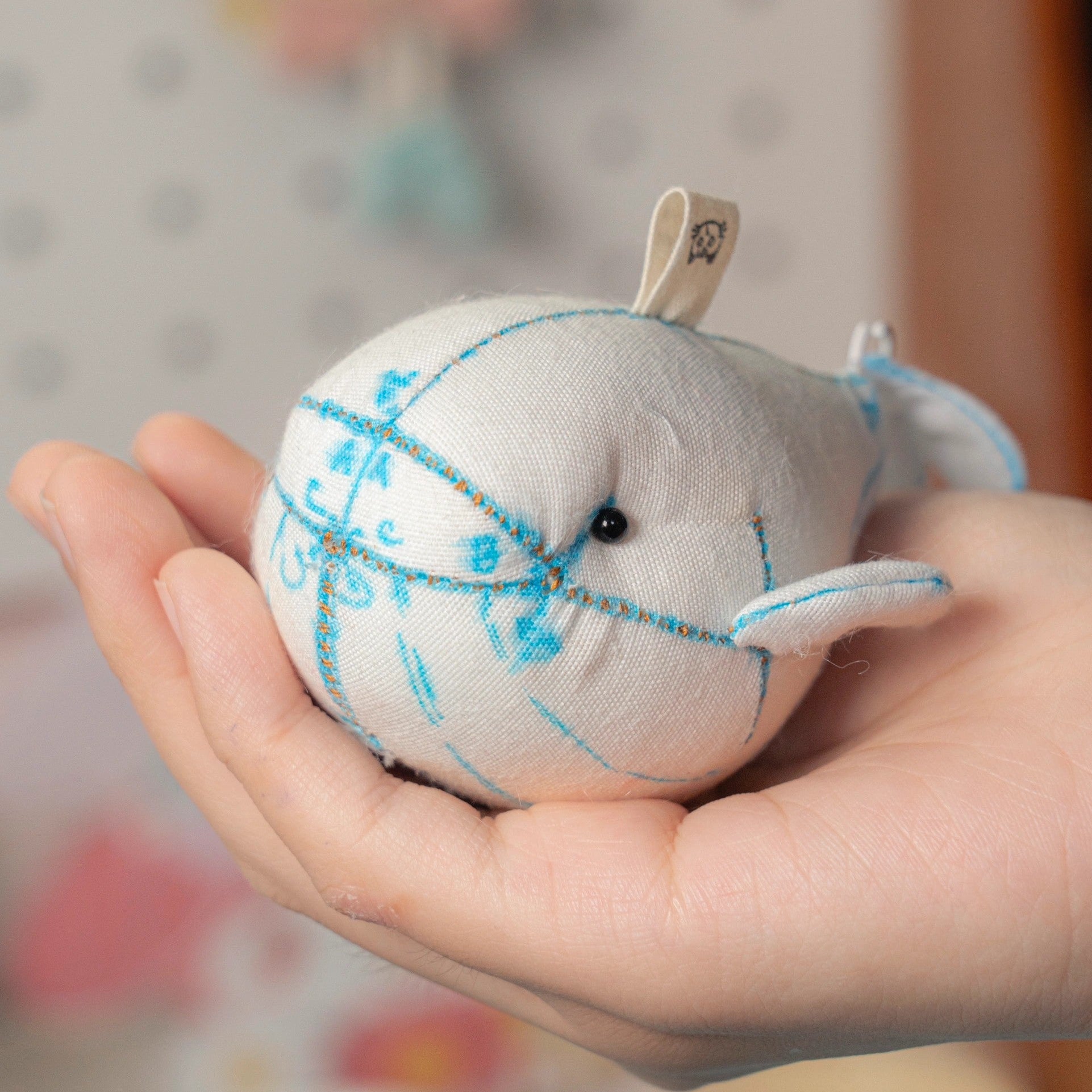 Hand holding a small plush whale toy with blue stitching against a blurred background