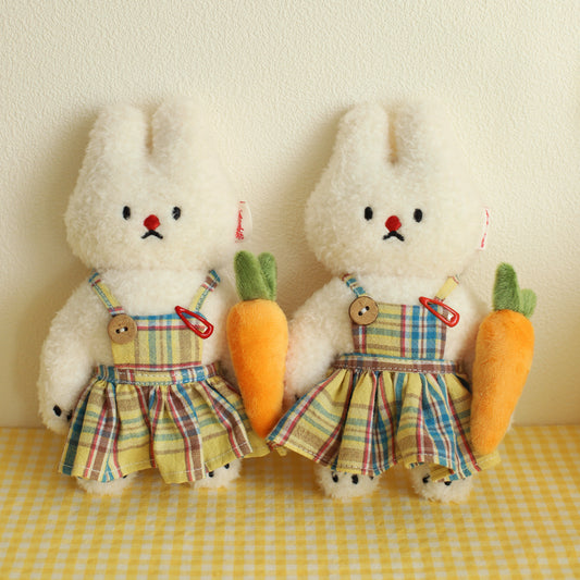Two plush bunnies in plaid dresses holding carrots on a yellow checkered tablecloth.