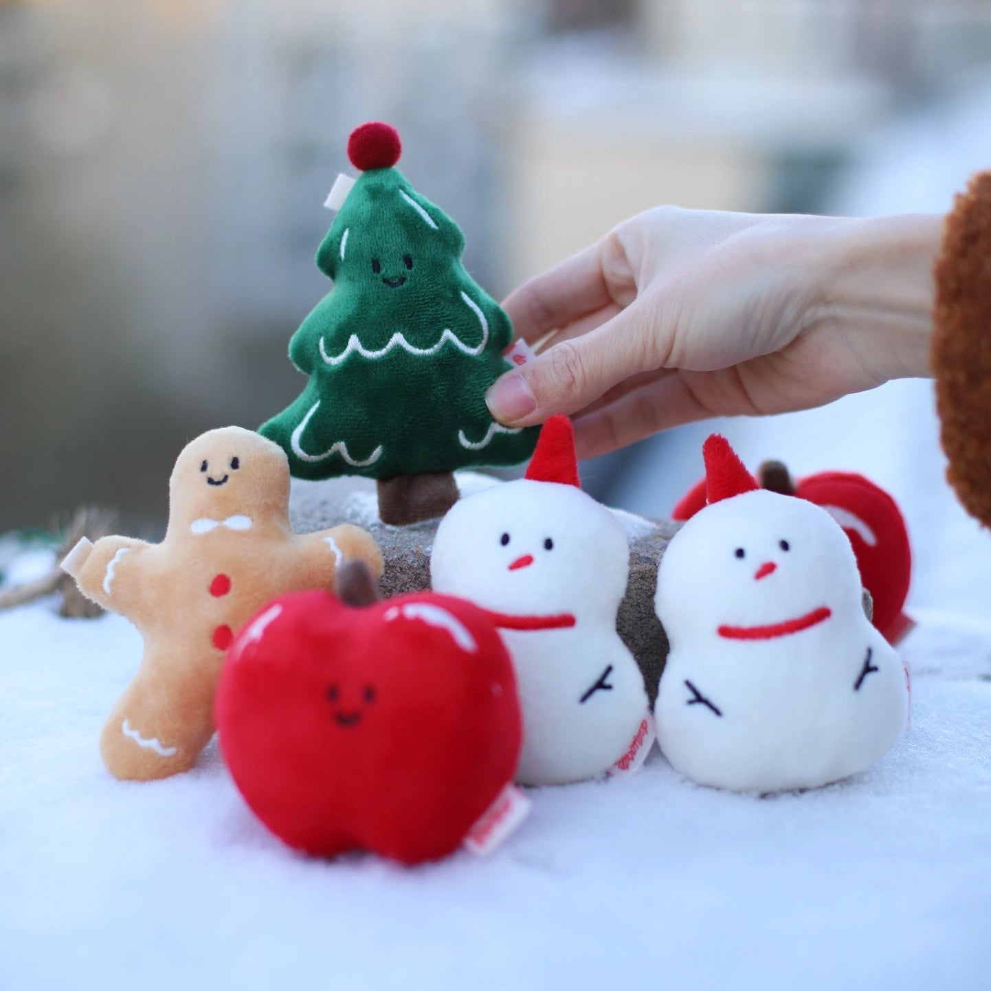 Hand holding a small green Christmas tree toy among other festive plush toys on a snowy background.