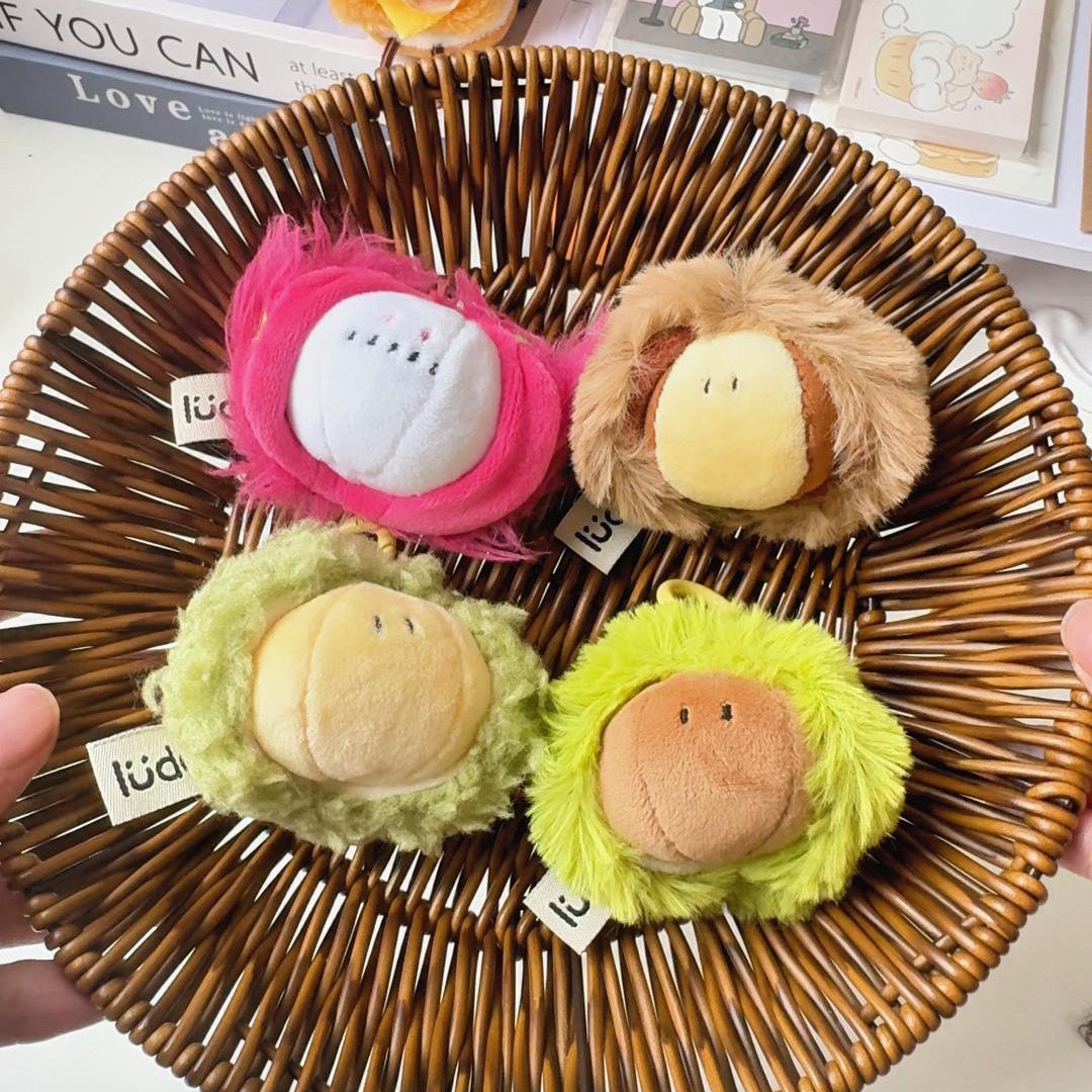 Four small plush character accessories arranged in a round woven brown basket, held by hands against a styled desk background with pens, stickers, and books.